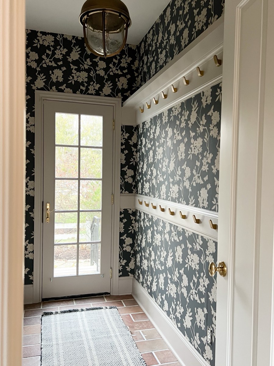 Dark floral wallpaper in a mudroom entryway with brass hooks, installed by Mancini Wallpapers